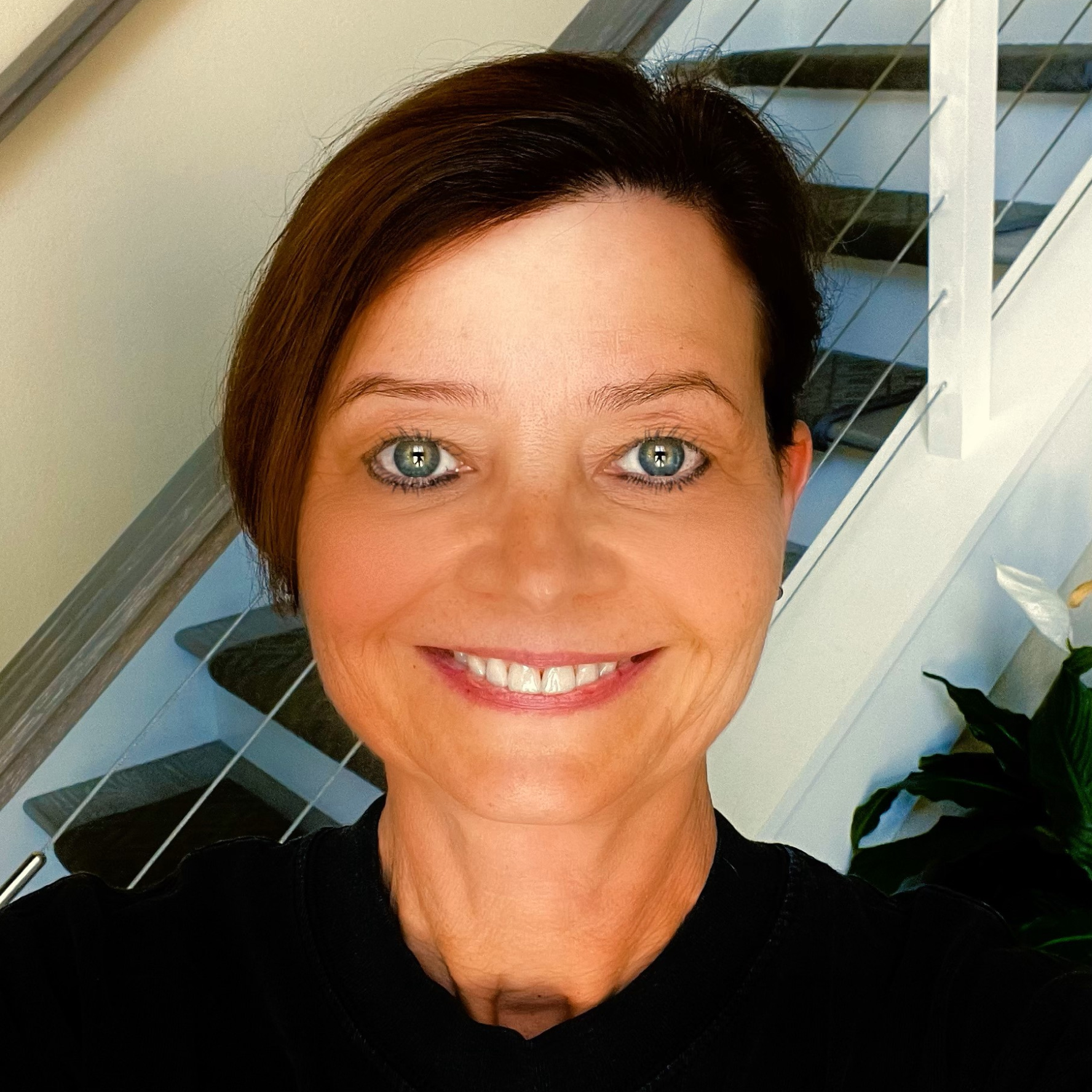 Laurie Kozbelt, also known as LK, a light-skinned woman with short, dark brown hair and green eyes, is smiling at the camera. She wears a black shirt and is standing beside a staircase.