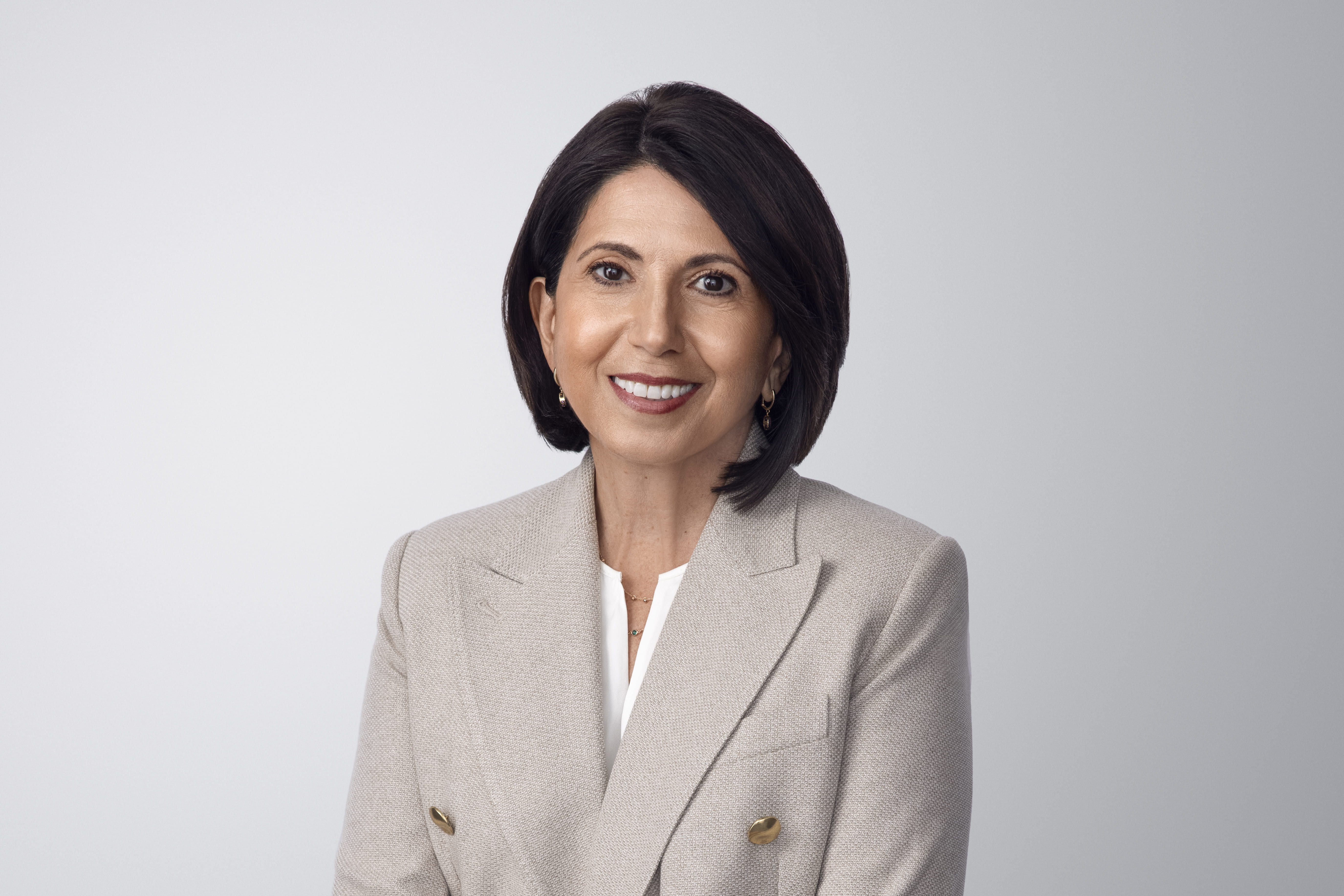 Fotografía de Silvana Battaglia. Silvana, una persona de piel clara con cabello castaño y ojos marrones, lleva aretes y sonríe a la cámara. Lleva una chaqueta gris sobre una camisa blanca y un collar. Está frente a un fondo gris claro sin entornos.