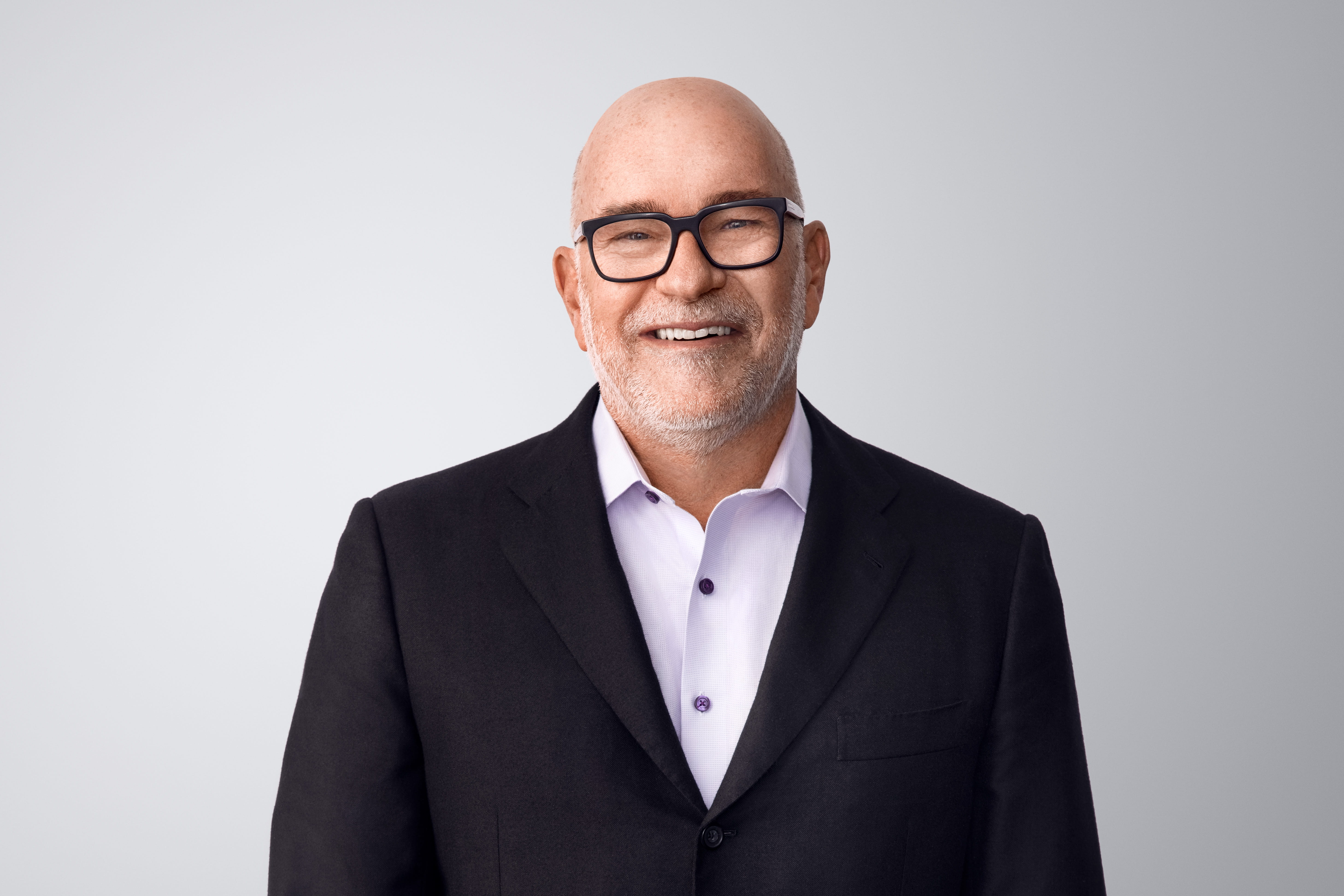Photograph of Steven H. Collis. Steven, a light-skinned person with dark framed eyeglasses and a short gray beard, is smiling at the camera. He wears a black suit jacket over a light purple shirt. He is in front of a light gray background with no surroundings.