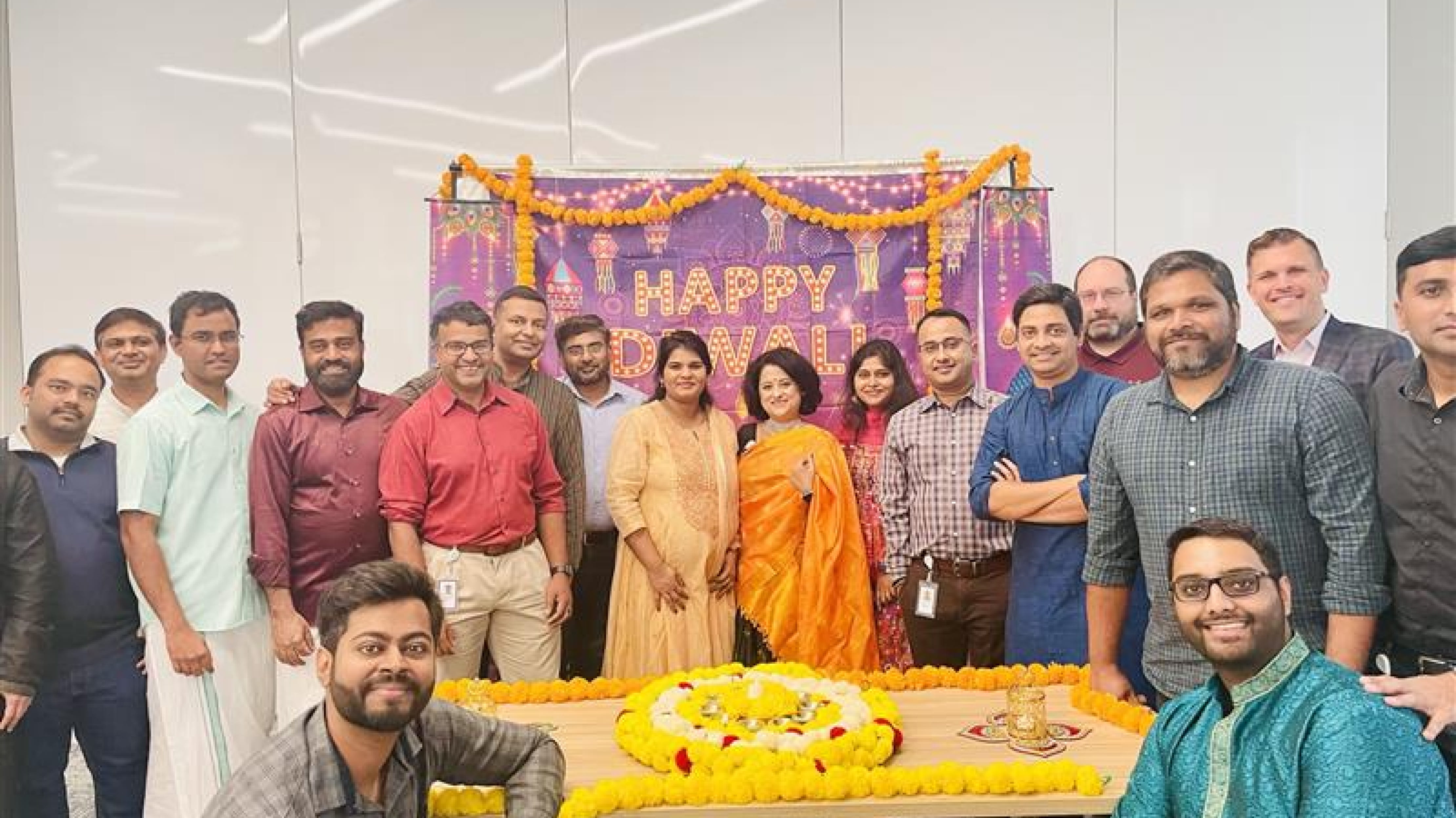 Un groupe de 18 employés de Cencora dans un bureau se tient devant une grande pancarte violette et or accrochée au mur sur laquelle on peut lire « Joyeux Diwali ».