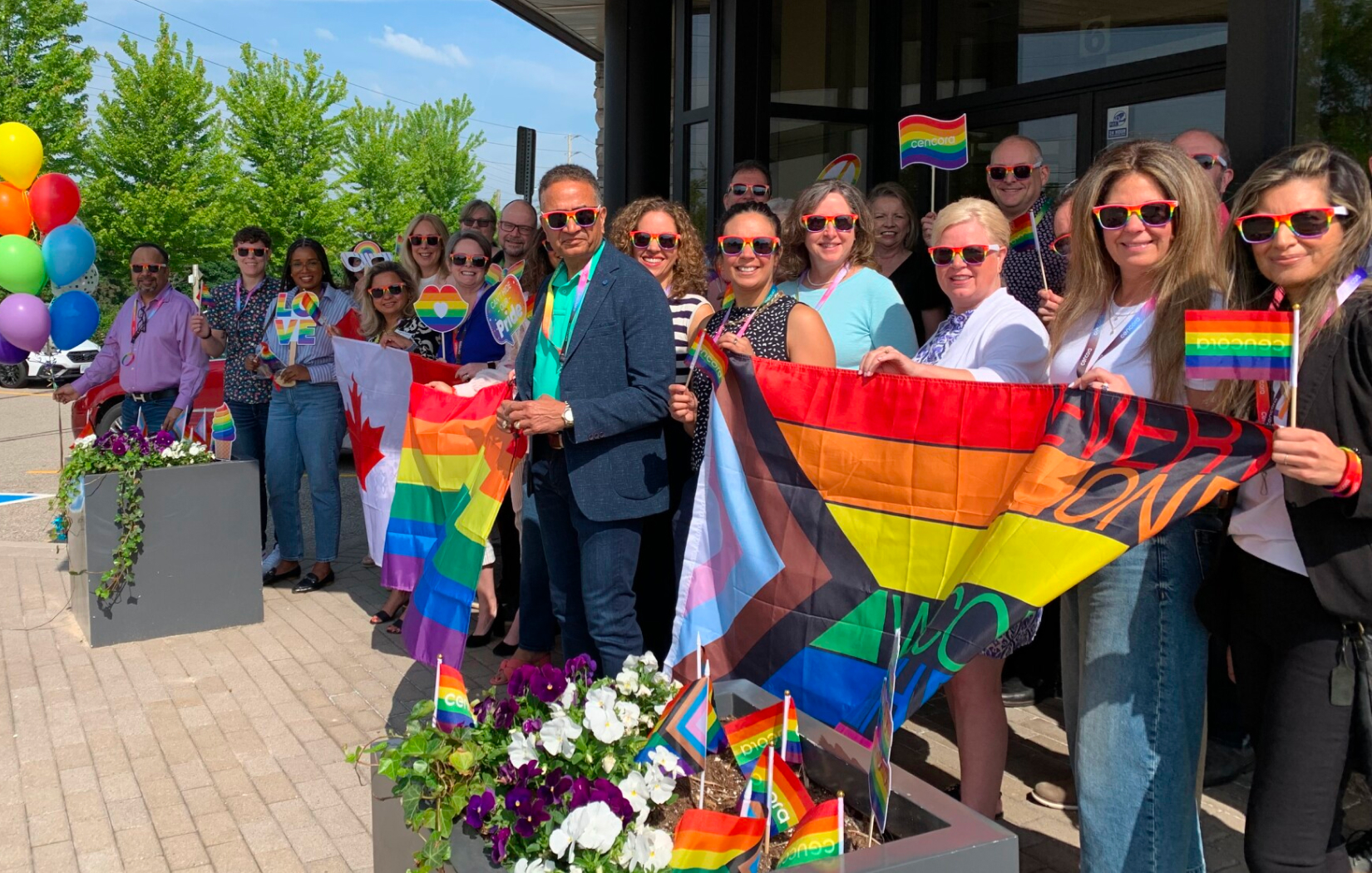A large group of Cencora team members and leaders celebrating Pride Month 2024 with a flag-raising event on the steps of their Conshohocken, Pennsylvania headquarters. They are dressed colorfully, including rainbow sunglasses.