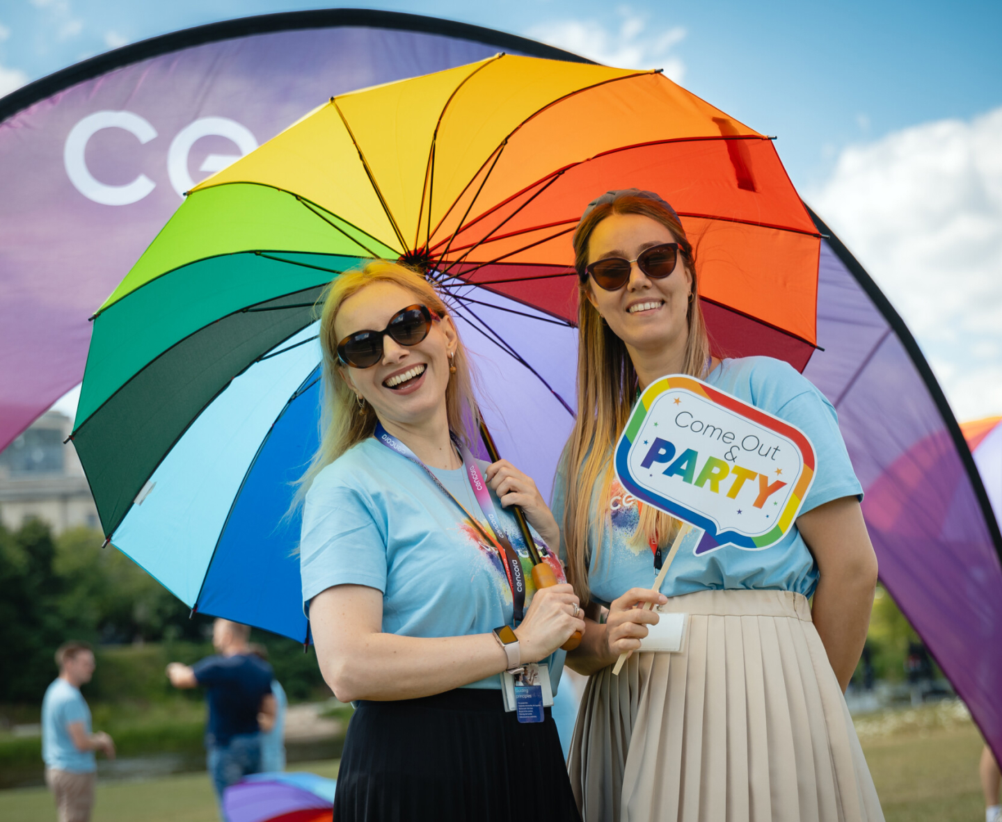 Two people standing under a large rainbow umbrella outside of the Cencora headquarters in Conshohocken, PA. One holds up the umbrella while the other holds a colorful sign that reads, "Come out and party."