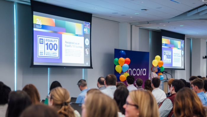 Employees gather in the Cencora office to watch a presentation. A person stands at a podium while two projection screens on either side display the Human Rights Campaign Foundation’s 2023-2024 Corporate Equality Index logo.