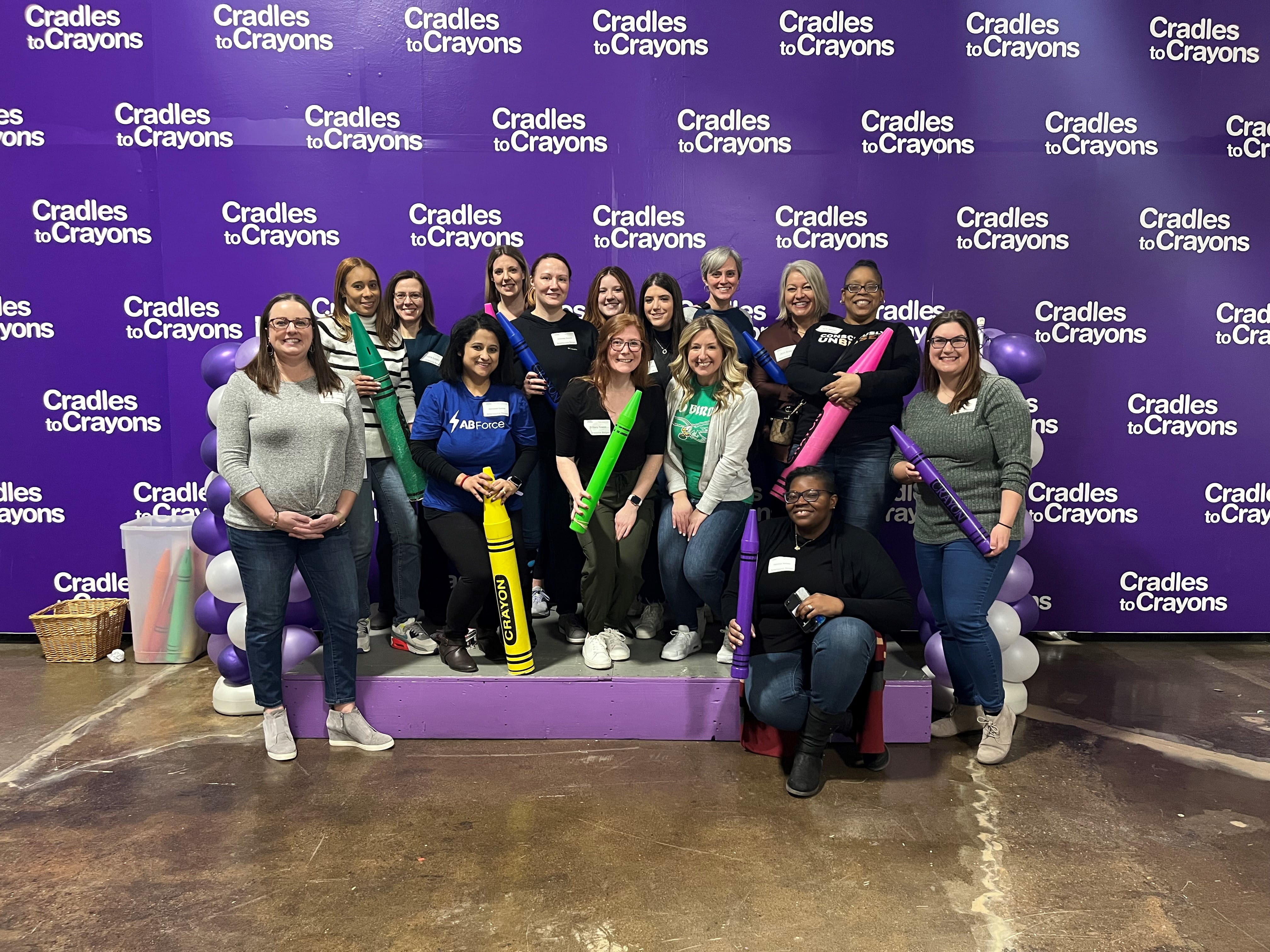 15 mujeres sonríen mientras sostienen grandes lápices de colores en un evento benéfico Cradles for Crayons. 