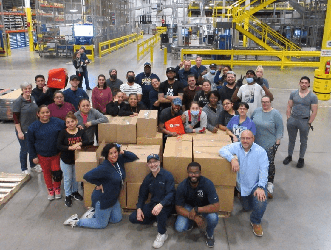 Des dizaines de personnes diverses posent pour une photo dans un entrepôt alors qu’elles se rassemblent autour d’une pile de grandes boîtes contenant des dons de bienfaisance.
