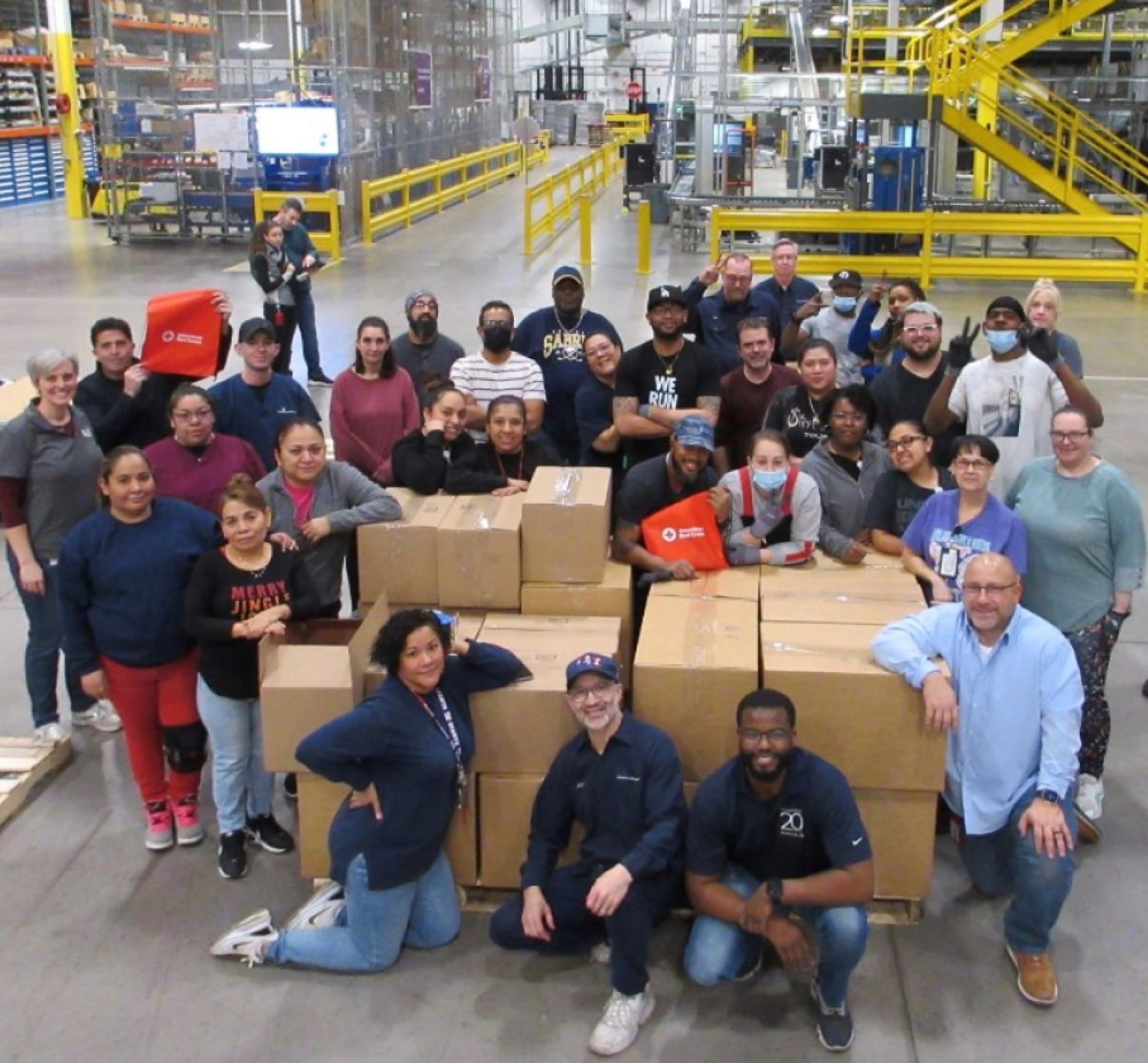 Des dizaines de personnes diverses posent pour une photo dans un entrepôt alors qu’elles se rassemblent autour d’une pile de grandes boîtes contenant des dons de bienfaisance.