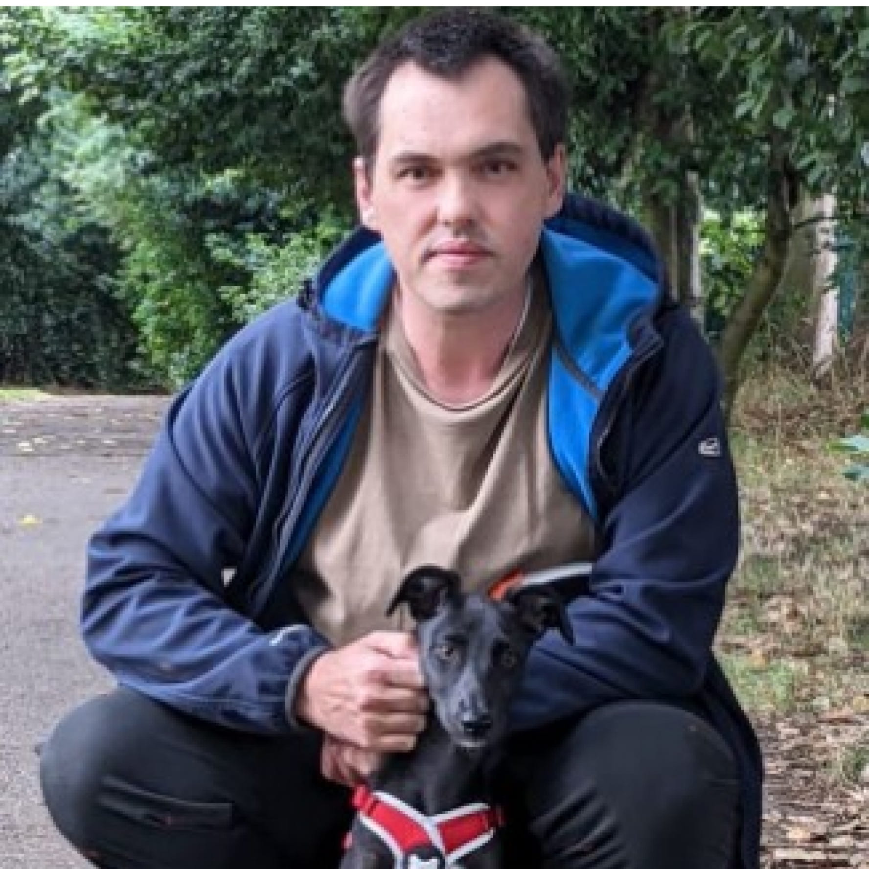David Steel arrodillado detrás de un pequeño perro negro. David está afuera en un sendero entre unos árboles y viste una sudadera con capucha azul marino con cremallera y una camiseta marrón.