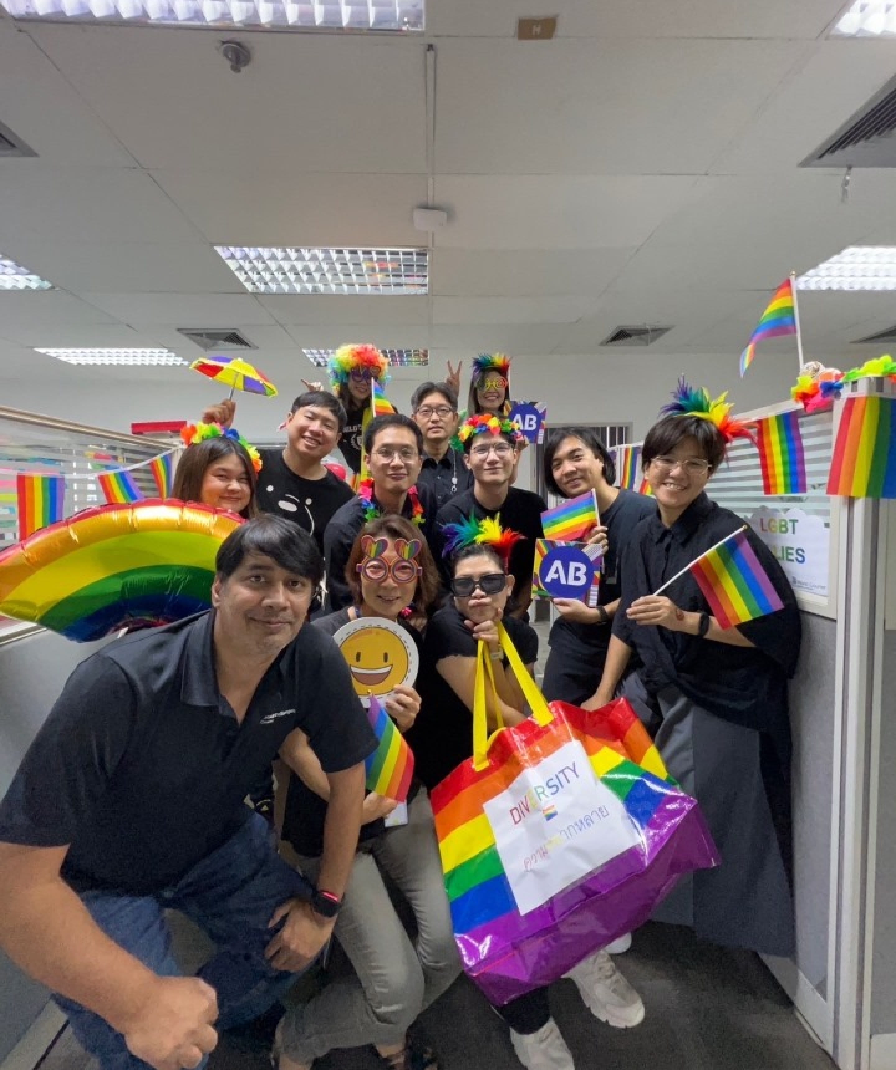 Un groupe d’employés tenant une variété de drapeaux arc-en-ciel, de ballons, de lunettes, de bandeaux et plus encore alors qu’ils posent parmi les cabines d’un bureau.