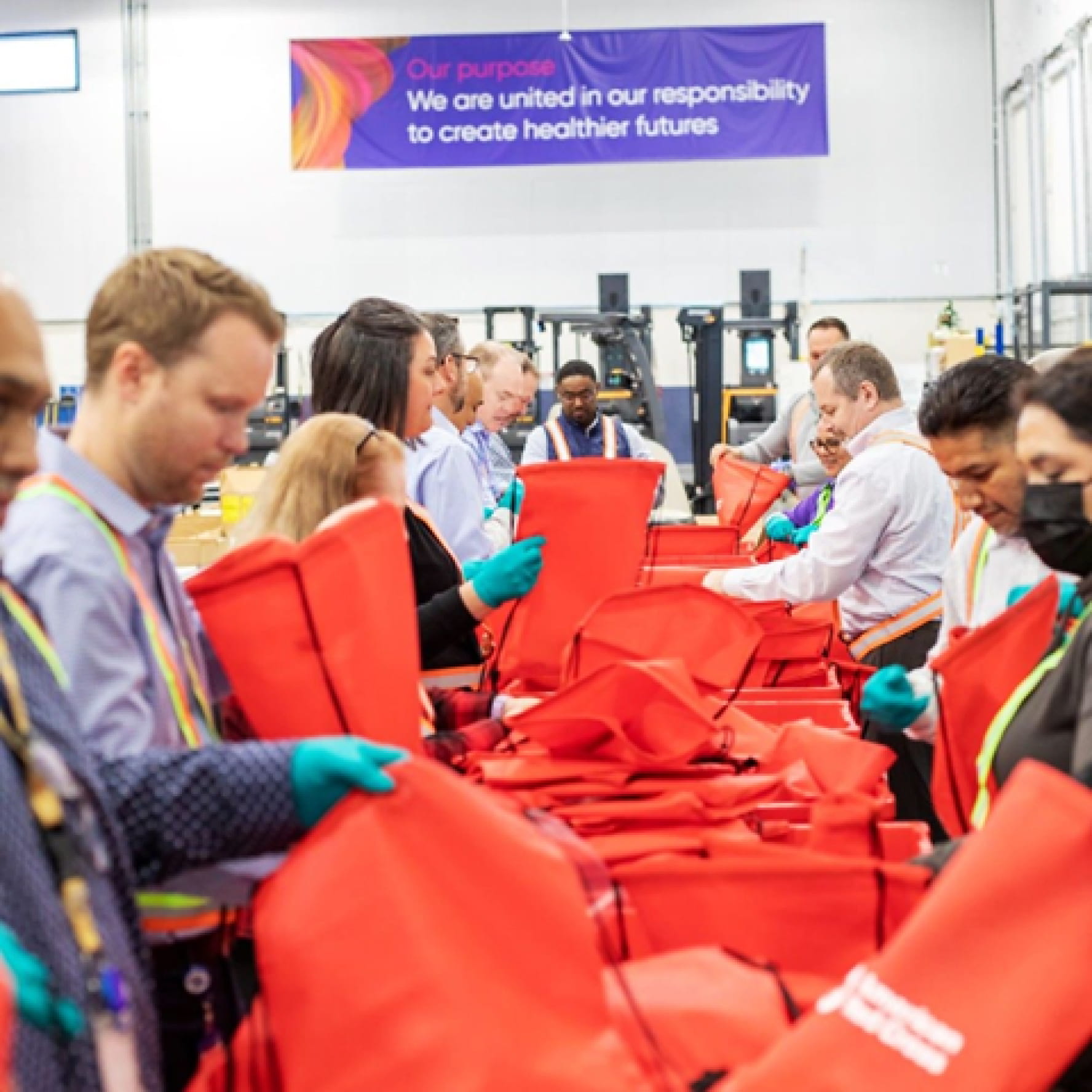 Una docena de voluntarios de Cencora empaquetando suministros de emergencia en bolsas de lona roja dentro de un almacén. Una pancarta en la pared detrás de ellos dice: "Nuestro propósito. Nos une la responsabilidad de crear futuros más saludables".