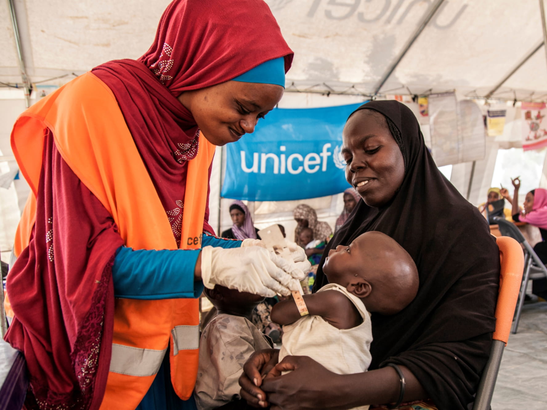 Una persona de piel oscura con un hiyab rojo y un chaleco naranja de Unicef colocando una pulsera a un pequeño niño de piel oscura dentro de un refugio con dosel blanco. Una tercera persona que lleva un hiyab negro sostiene al niño. Una pancarta azul de Unicef cuelga detrás de ellos.