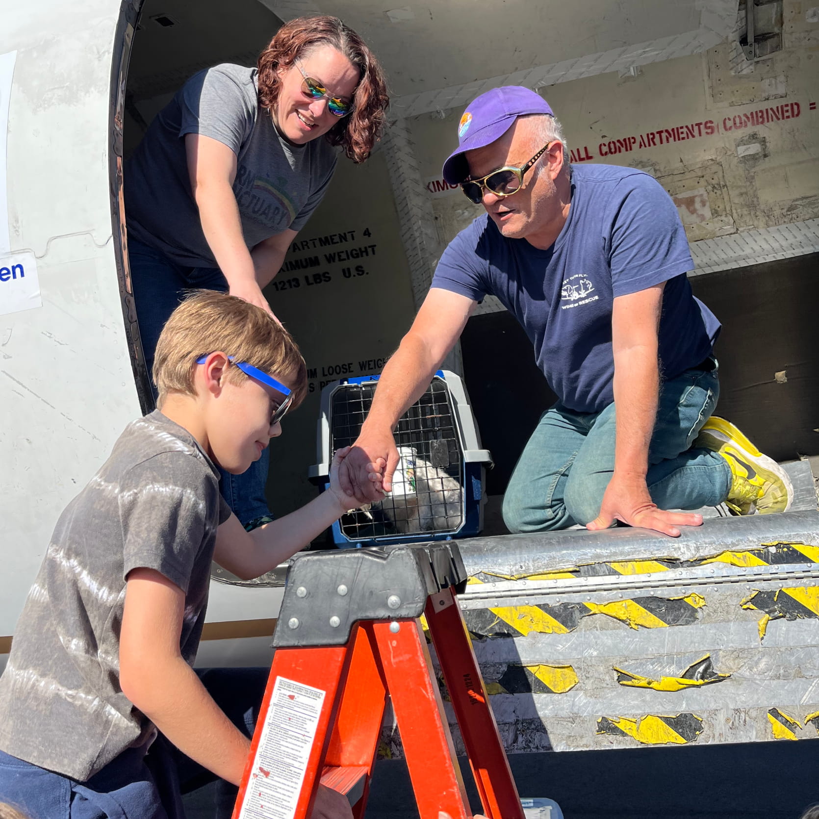 Un hombre, una mujer y un niño que trabajan con la organización benéfica Wings of Rescue se encuentran en un avión de carga con una jaula para mascotas visible en el fondo. 