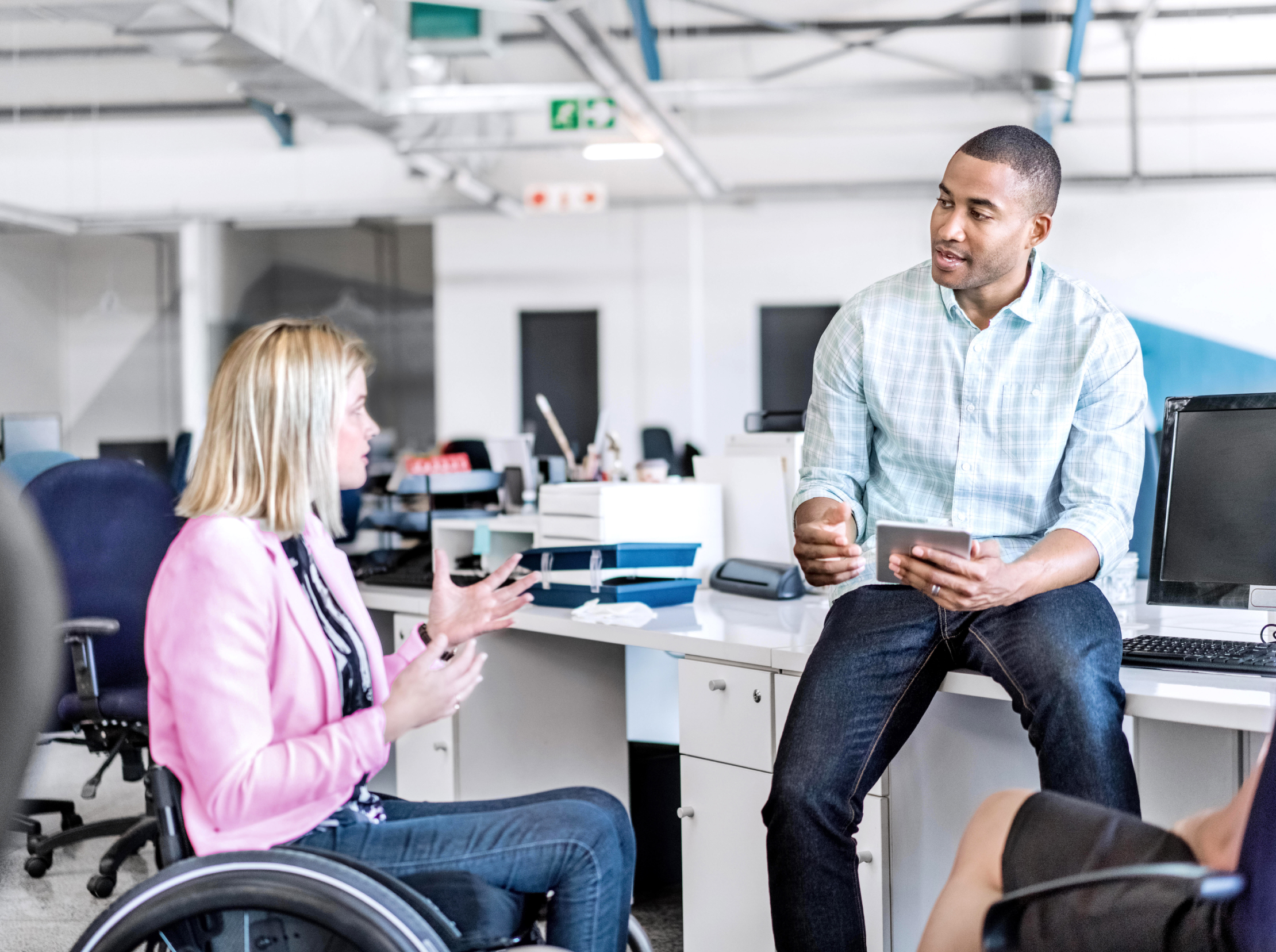 Zwei Kollegen (eine hellhäutige Person mit schulterlangen Haaren, die im Rollstuhl sitzt, und eine dunkelhäutige Person mit kurzen Haaren, die auf der Kante eines Schreibtischs sitzt) in Business-Casual-Kleidung unterhalten sich in ihrem Büro.