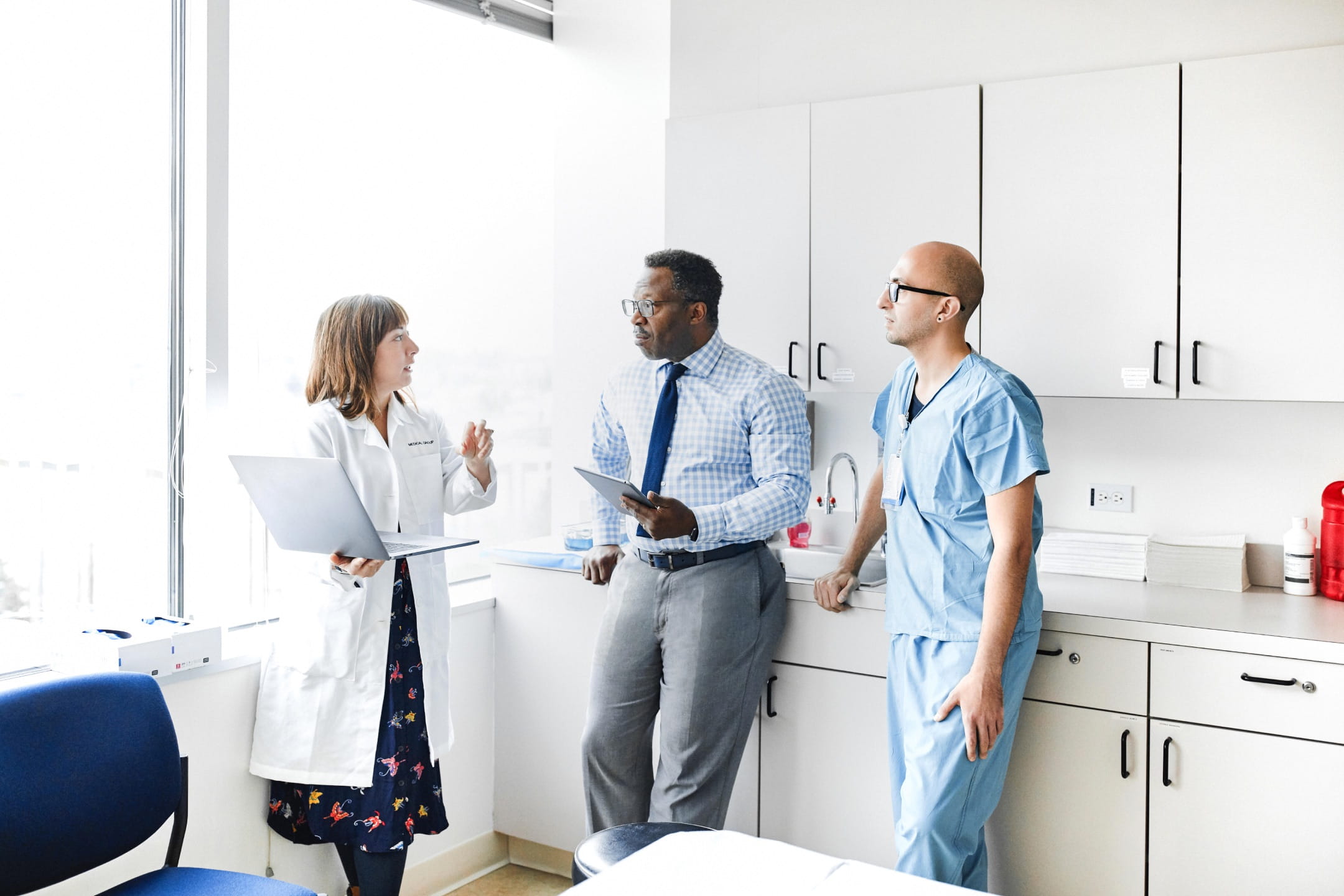 Tres profesionales médicos (un médico de piel clara de pie sosteniendo una computadora portátil, una persona de piel oscura con ropa de negocios sosteniendo una tableta y una enfermera de piel clara con bata) conversando en el consultorio de un médico.