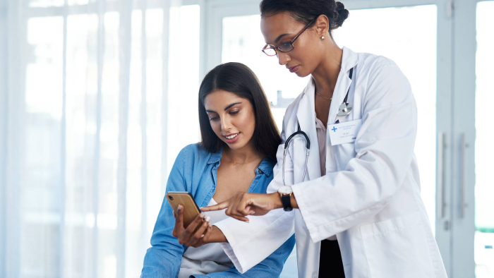 Ein Arzt zeigt einem Patienten Informationen auf einem Mobiltelefon in einem hellen Raum. Beide haben lange dunkle Haare und mittlere Haut. Der Profi trägt einen weißen Laborkittel, Ohrringe und ein Stethoskop. Der Patient trägt ein Jeans-Langarmhemd mit einem weißen Hemd darunter.