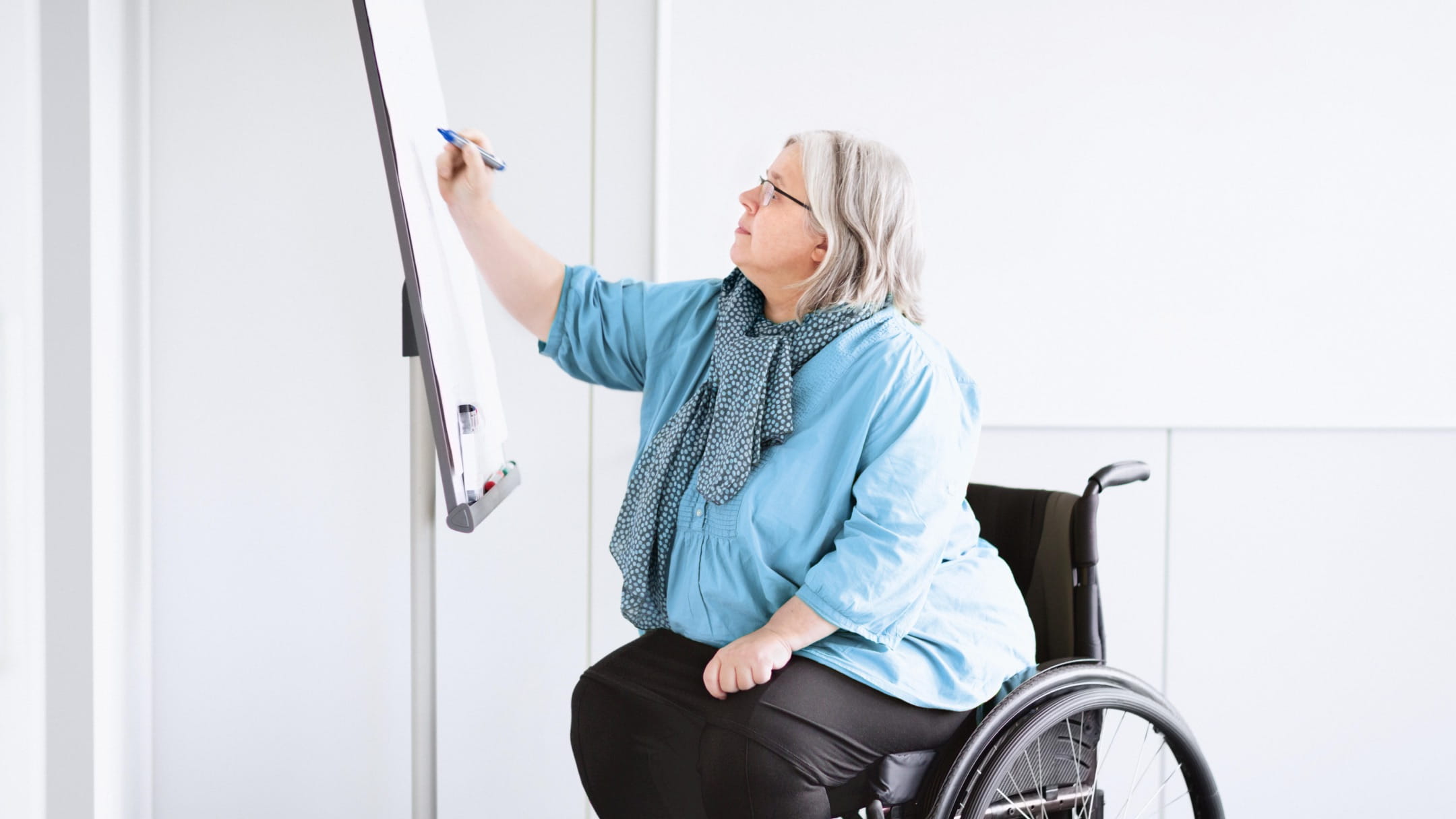 Une personne en fauteuil roulant écrit sur un tableau blanc dans une salle de réunion lumineuse. Ils ont la peau claire, les cheveux gris jusqu’aux épaules et portent des vêtements décontractés avec des lunettes et une écharpe bleue.