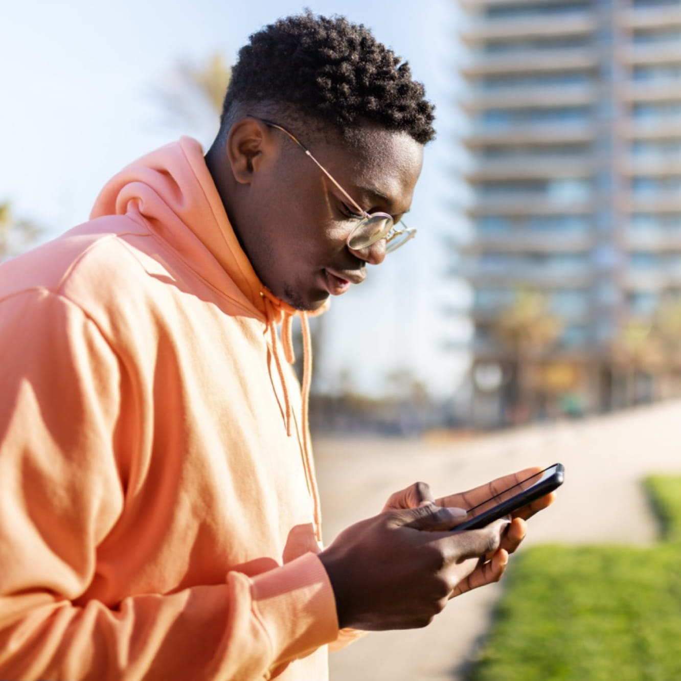 Un jeune adulte à la peau foncée regarde son téléphone portable. Ils ont les cheveux courts et portent un sweat-shirt à capuche orange et des lunettes. Ils sont à l’extérieur devant un grand bâtiment. 