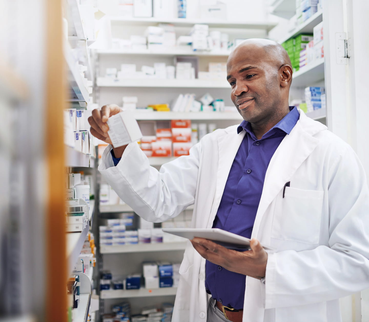 A dark-skinned, bald pharmacist examining a medication box in the stock room of a pharmacy, surrounded by shelves of medication. They are wearing a purple collared shirt underneath a white lab coat.