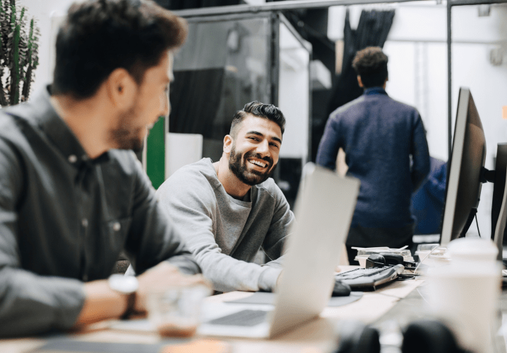 Two young men are working on laptops and casually chatting with each other in an office. 