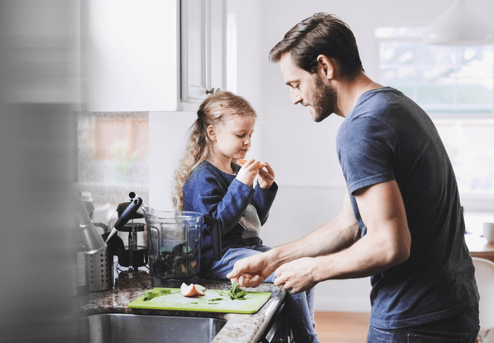 Una giovane ragazza siede su un bancone della cucina mangiando frutta mentre suo padre pulisce l'area di preparazione del cibo.