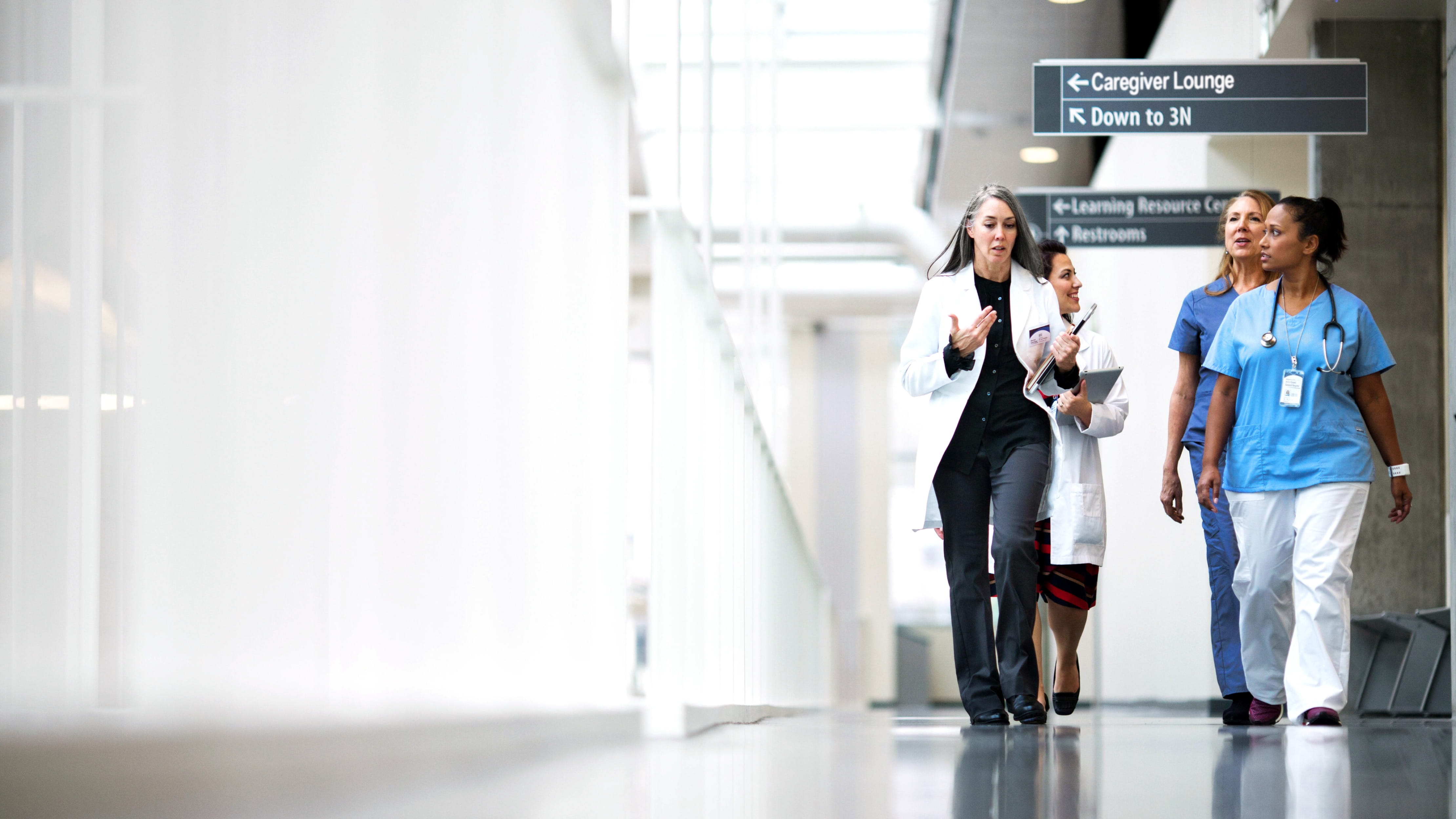 Quatre professionnels de la santé marchant dans un couloir tout en discutant. Deux d’entre eux portent des blouses blanches et portent des appareils de table. Les deux autres personnes portent des blouses.