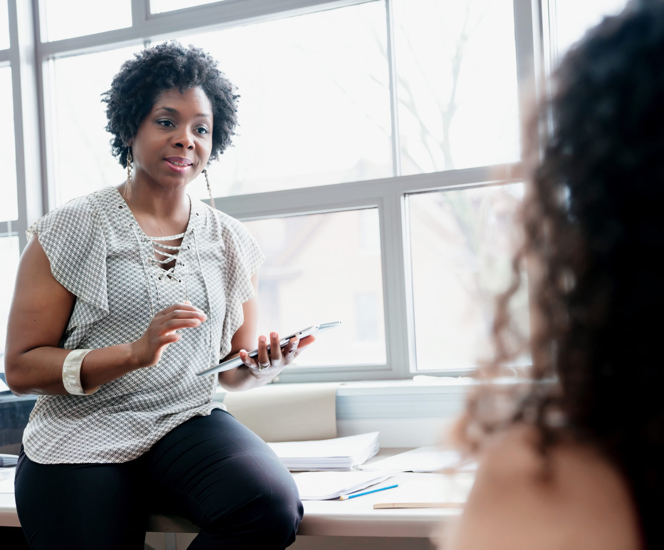 Une personne à la peau foncée et aux cheveux courts, tenant une tablette, s’appuie sur un bureau tout en discutant avec une personne à la peau claire assise en face d’elle.