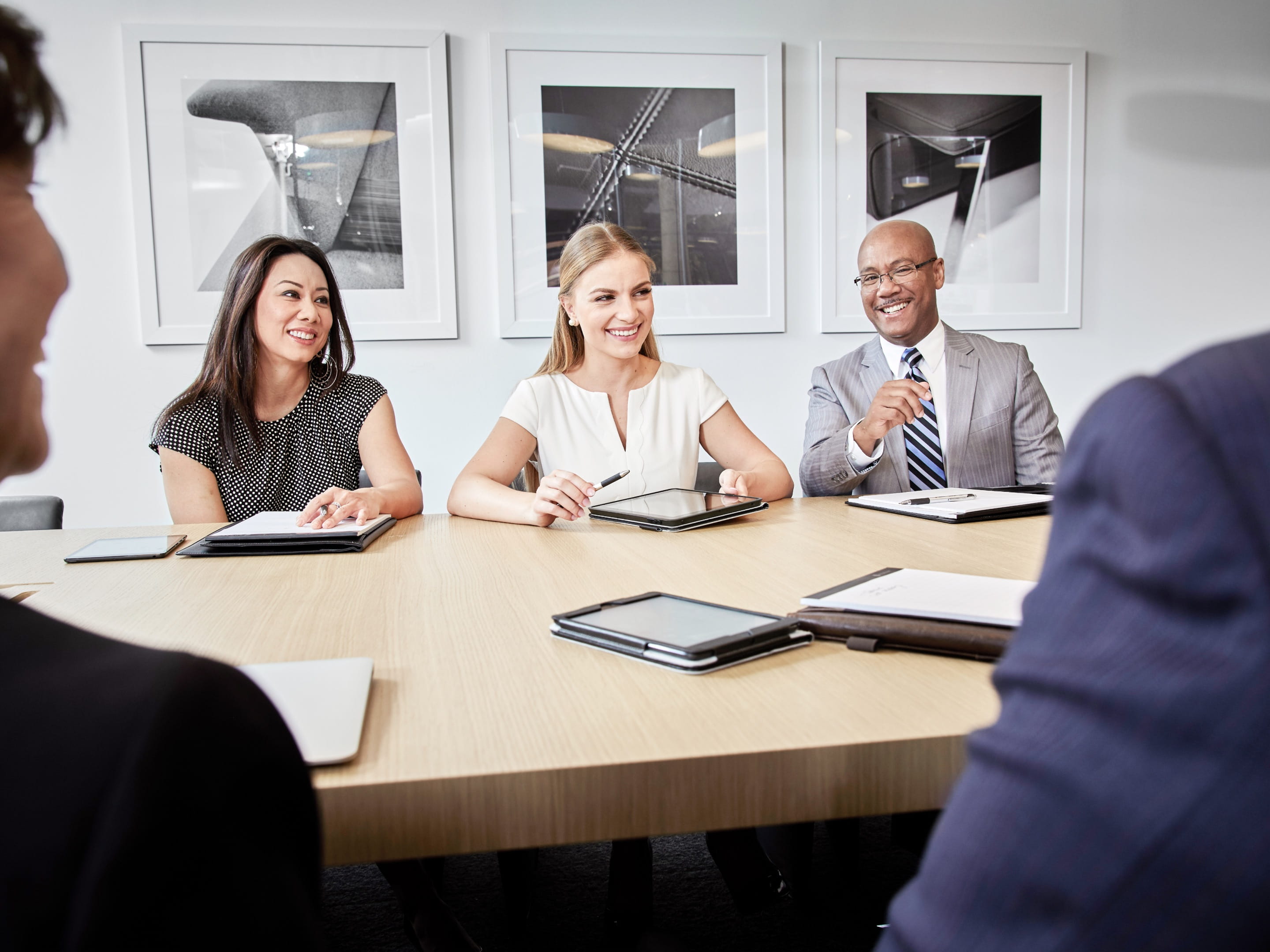 Un groupe diversifié d’employés vêtus de vêtements d’affaires assis à une table dans une salle de réunion. Des tablettes, des blocs-notes et des stylos sont posés sur la table devant eux.