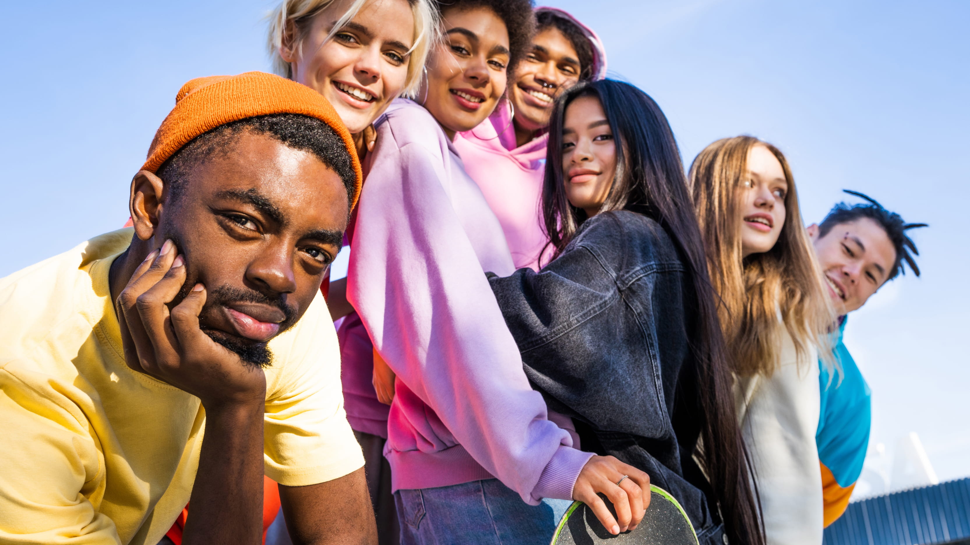 A diverse group of seven young adults posing in a line for a photo. One sits and the rest stand. One glances off in the distance while the rest look at the camera. Most have a slight or full smile. They wear colorful clothes and a clear blue sky is visible above them.  