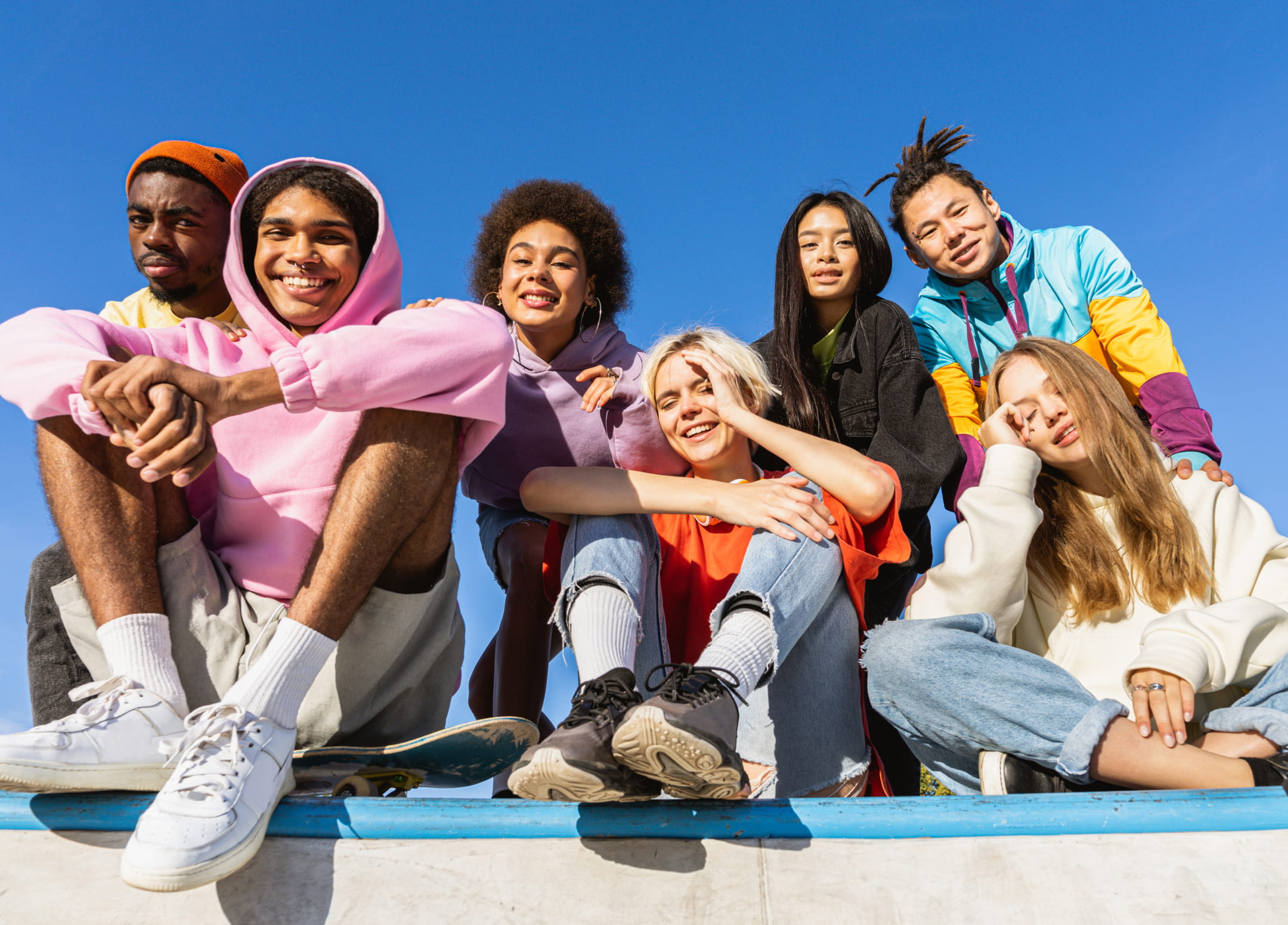 A diverse group of seven young adults hanging out together on a cement ledge. The three in front are sitting while the rest kneel or stand behind them. Most of them are smiling. They each wear colorful clothes and a clear blue sky is visible above them. 