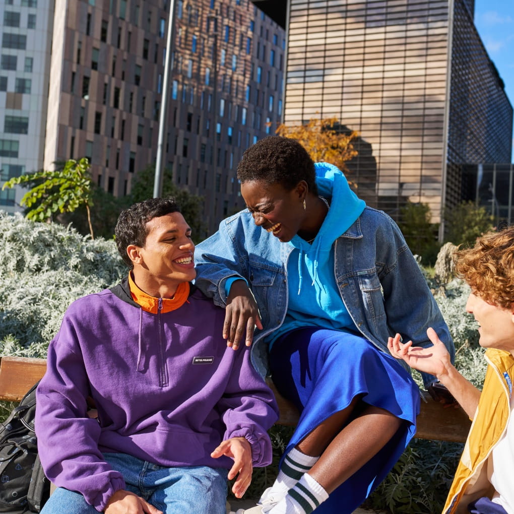 Tres jóvenes, vestidos con ropa deportiva informal, riendo y sonriendo sentados en un banco en un entorno urbano.