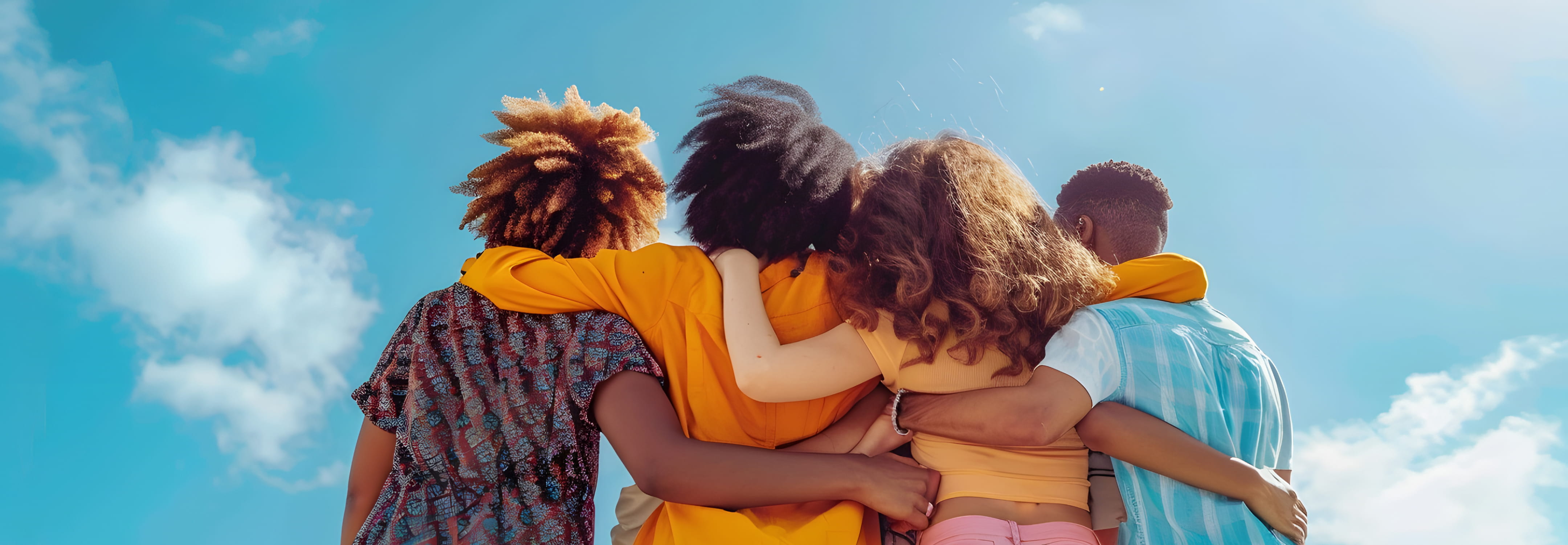 A diverse group of four young adults embracing each other side-by-side with hugs. They all wear colorful clothes and face away from the camera. A blue sky with a few clouds is visible overhead.