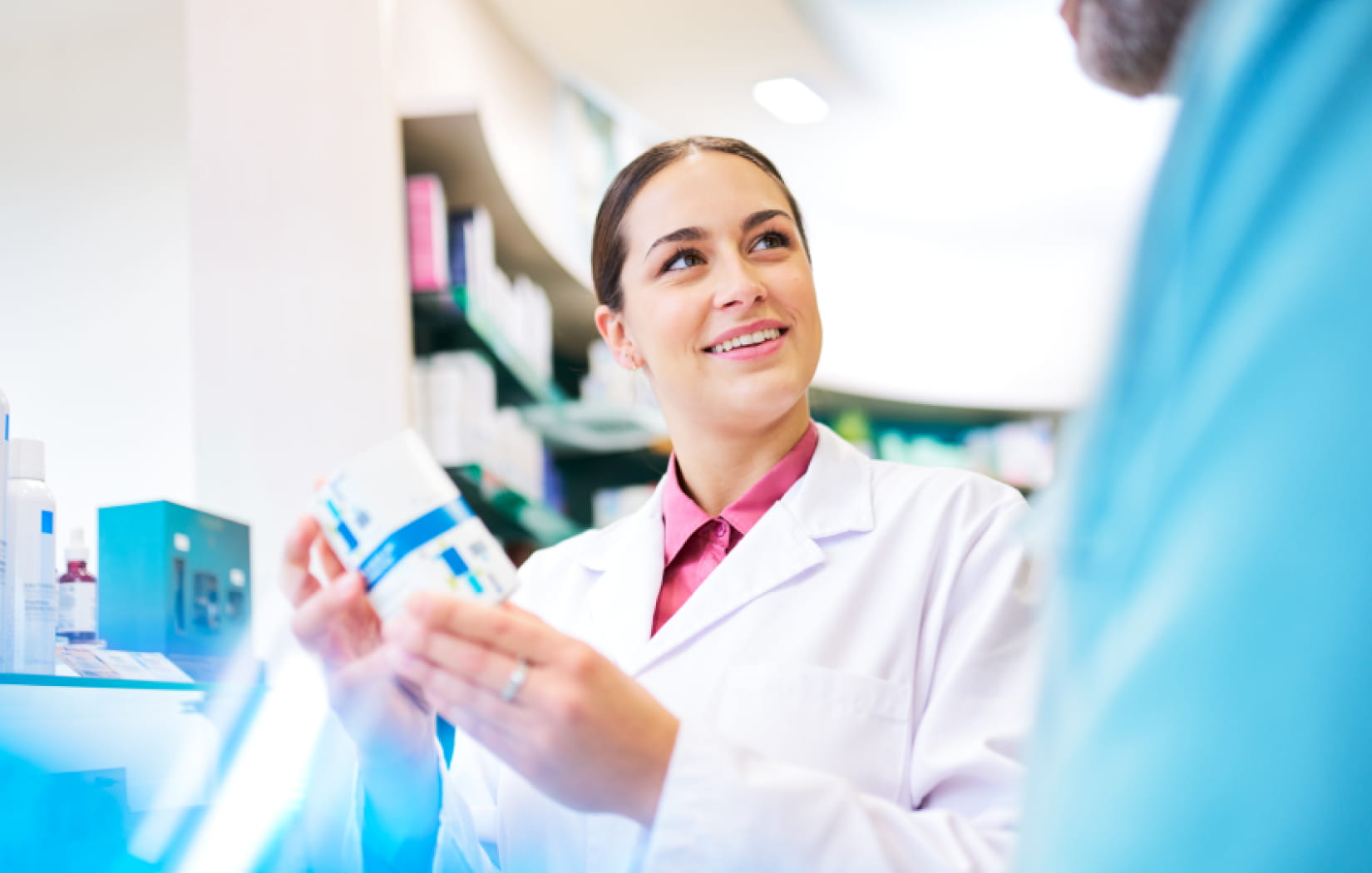 A light-skinned pharmacist holding up a box of medication to show another person who is mostly out of frame. They wear a pink collared shirt beneath a white lab coat. Shelves of medication are located behind them.