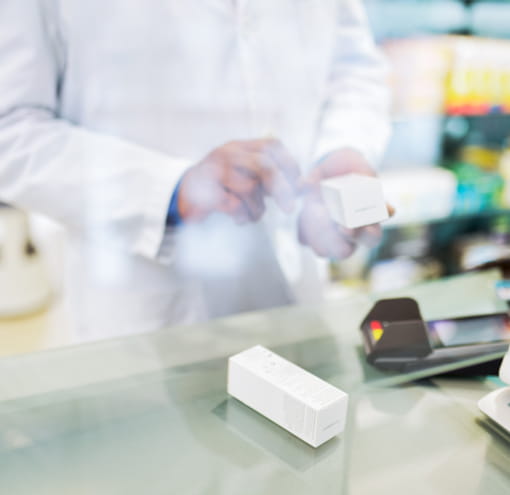 A pharmacist ringing up a customer’s purchase at a cash register. Only their hands are visible. They hold a small, white medication box and another sits on the counter.