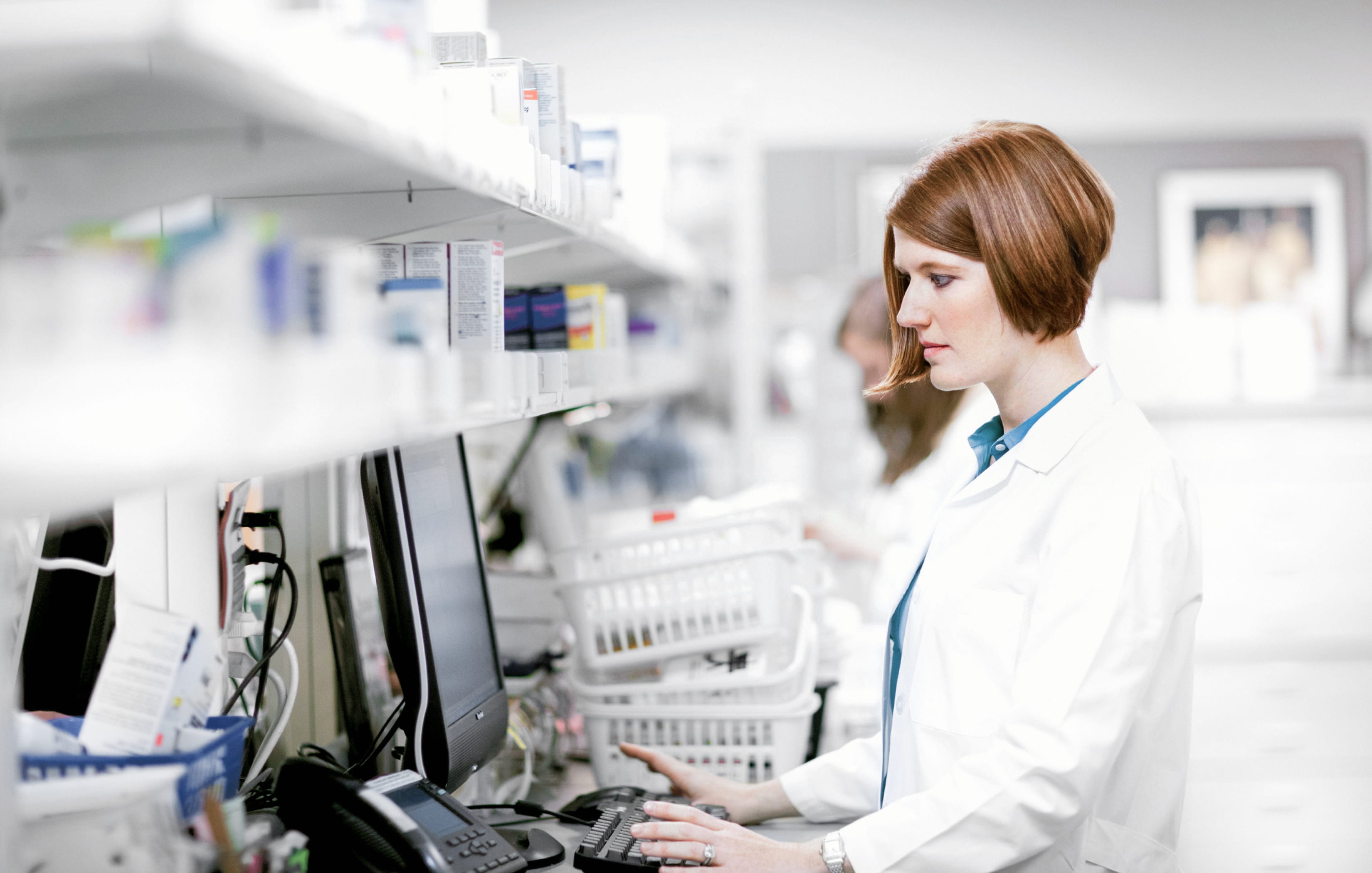 A pharmacist wearing a white lab coat standing in front of a desktop monitor fulfilling in order for a patient.