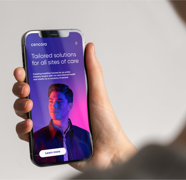 A hand holds a mobile phone with the new Cencora website showing in the browser screen. 