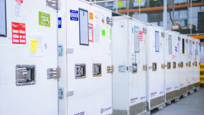 Eight Cencora shipping containers standing in a row in a warehouse. Each white container has various colorful stickers with words like, “refrigerator” and “controlled ambient.”