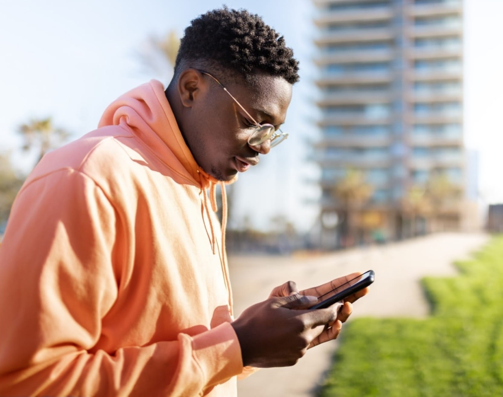 A dark-skinned young adult looking down at their cell phone. They have short hair and wear an orange hooded sweatshirt and glasses. They are outdoors in front of a large building. 