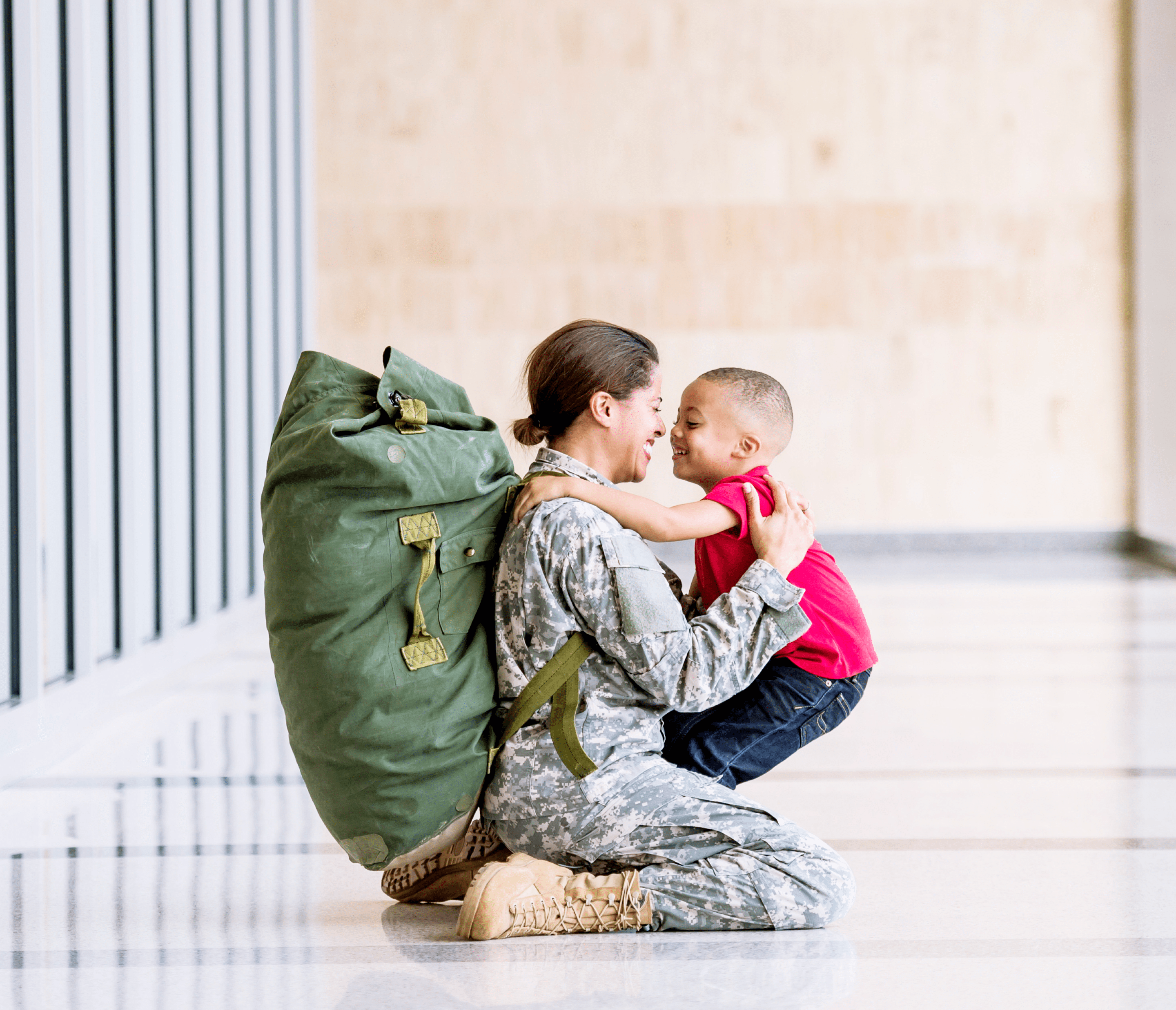 Un soldat à la peau moyenne assis sur le sol et tenant de près un enfant à la peau moyenne qui sourit. Le soldat porte un uniforme de camouflage et un grand sac à dos vert. L’enfant porte un t-shirt rouge et un jean bleu.