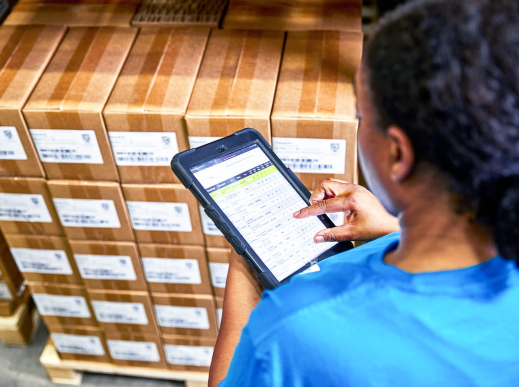 A warehouse worker wearing a blue t-shirt reviewing inventory data on a small tablet device standing in front of a pallet of boxes.