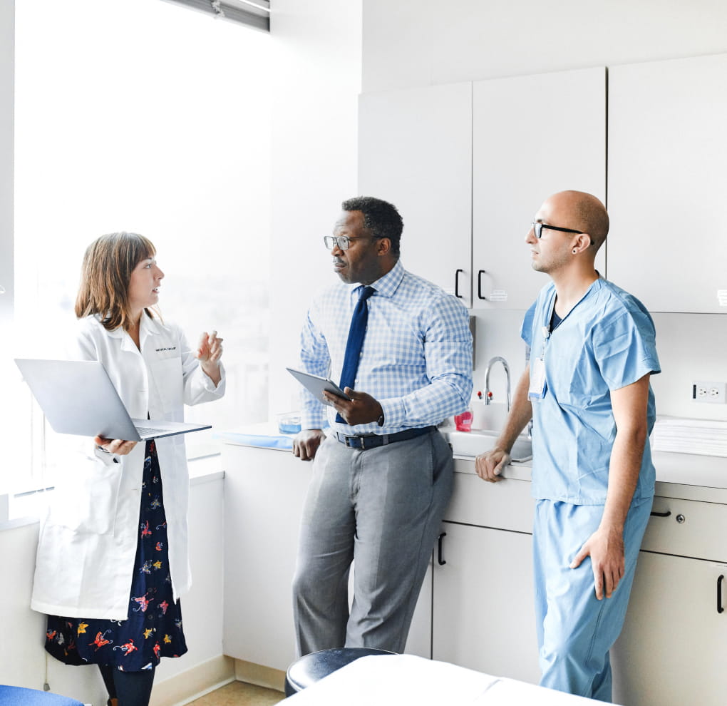Three medical professionals (a light-skinned doctor standing holding a laptop, a dark-skinned person in business clothing holding a tablet device, and a light-skinned nurse in scrubs) having a conversation in a doctor’s office.