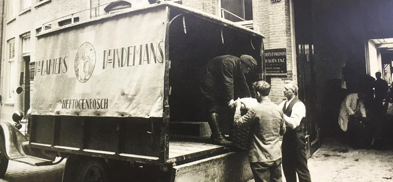  Historic photo showing workers loading goods onto a truck in a alley, with several people working in a warehouse in the background