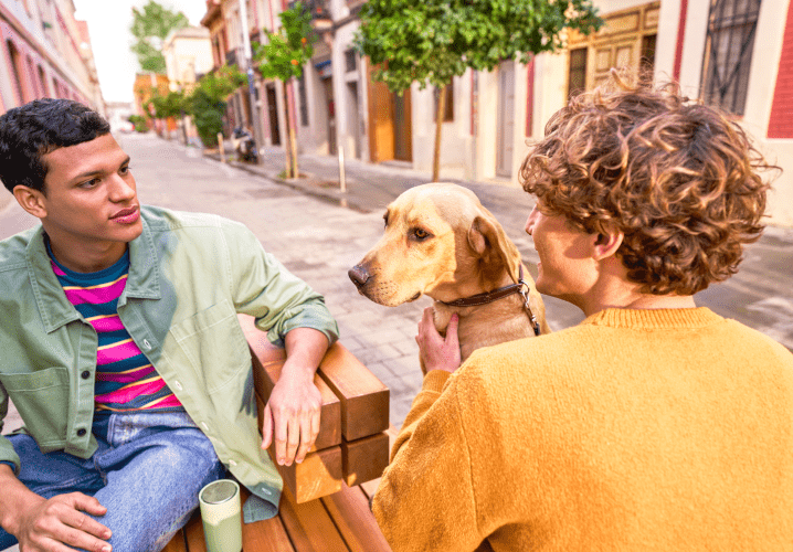 Deux jeunes hommes discutent sur un banc en plein air tout en caressant un chien.
