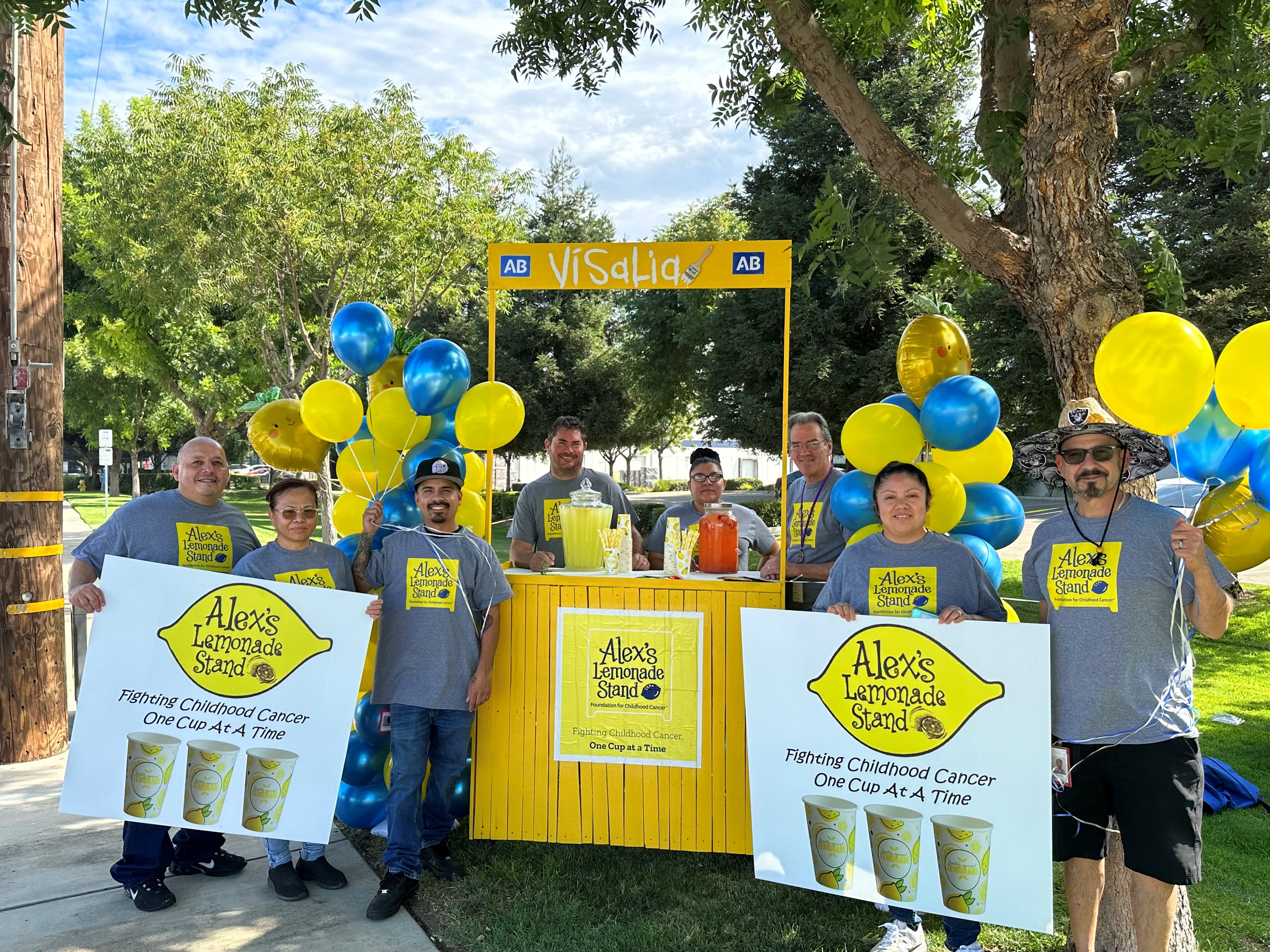 Eight Cencora volunteers posing outside a yellow lemonade stand with posters and T-shirts reading "Alex's Lemonade Stand."