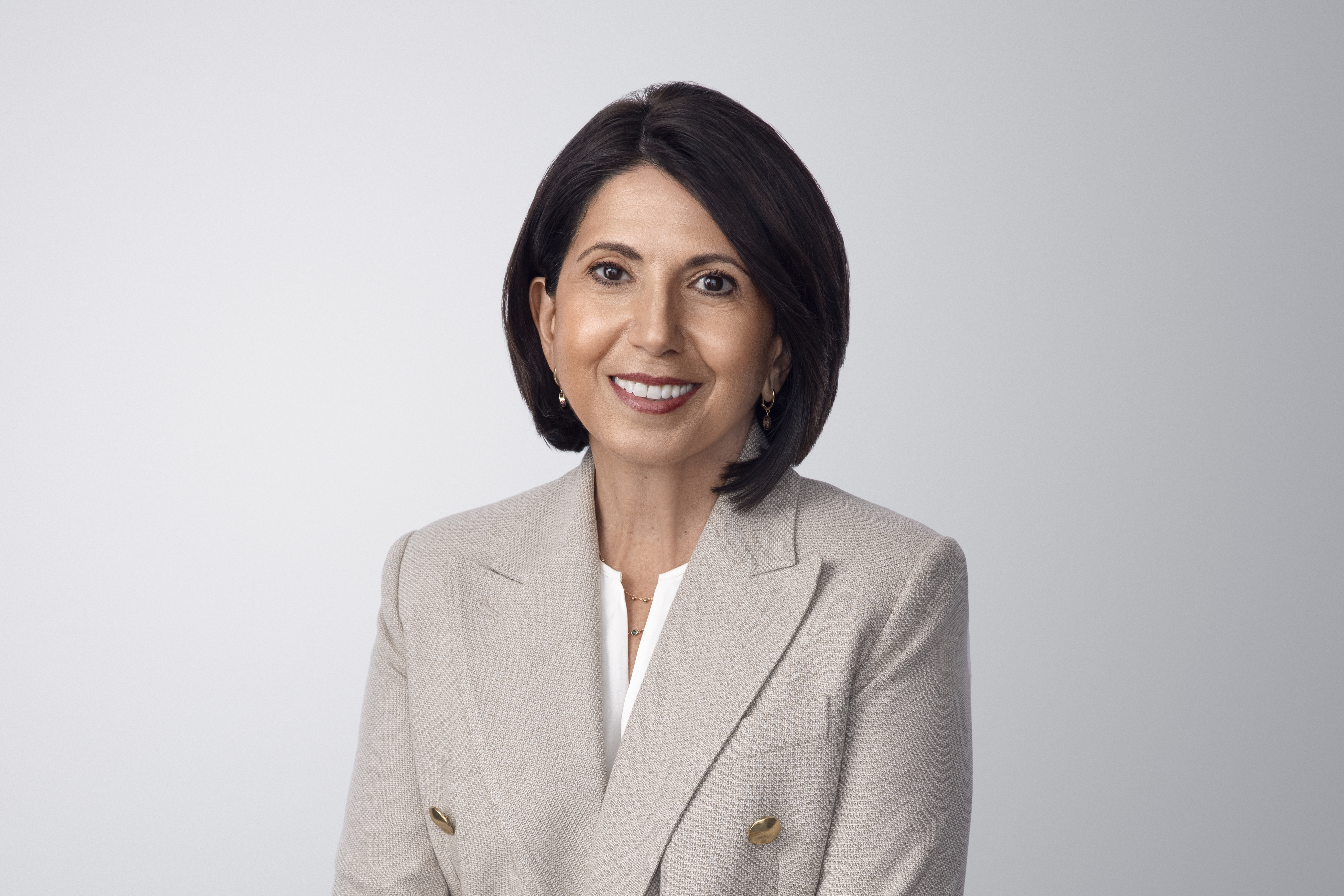 Fotografía de Silvana Battaglia. Silvana, una persona de piel clara con cabello castaño y ojos marrones, lleva aretes y sonríe a la cámara. Lleva una chaqueta gris sobre una camisa blanca y un collar. Está frente a un fondo gris claro sin entornos.